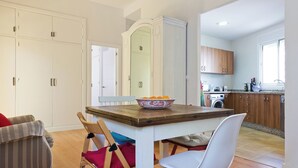 Une cuisine avec une table à manger, des chaises et un saladier rempli de fruits. On aperçoit un lave-linge et des meubles en bois à l’arrière-plan.