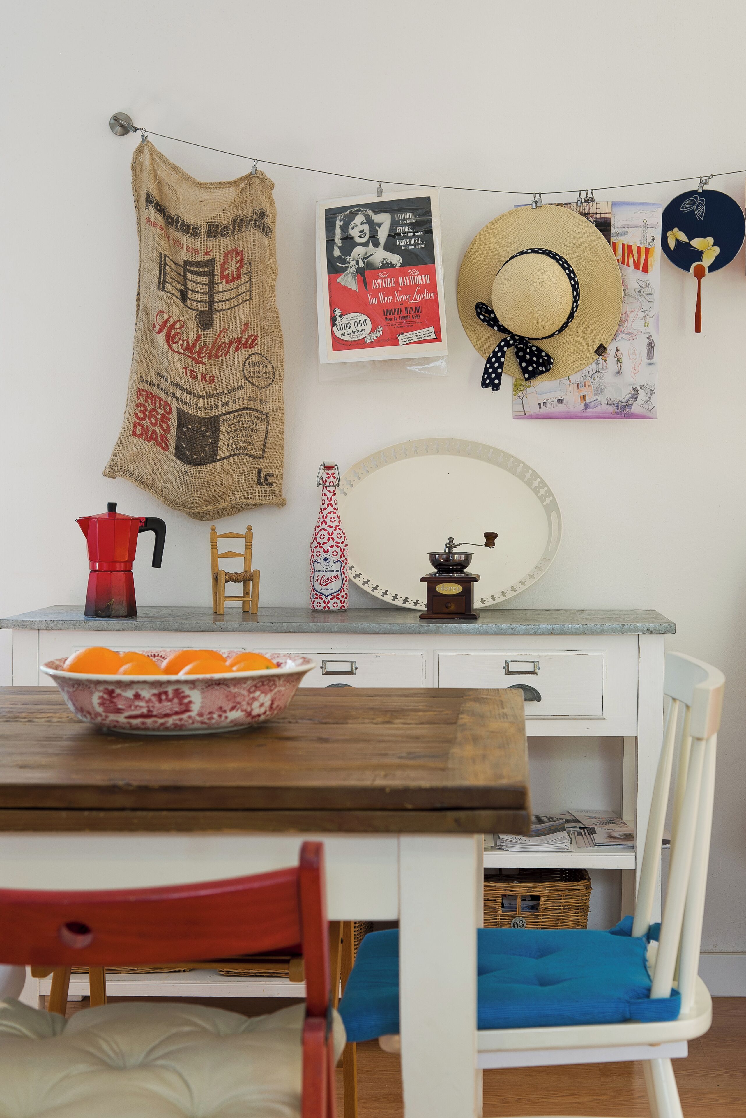 Una cocina con una mesa de madera, un cuenco de naranjas, una cafetera roja, una silla de madera, un armario blanco y un paño colgado.