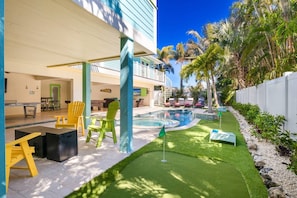Un área de piscina con un pequeño campo de práctica de golf, mobiliario exterior y un edificio con balcón.