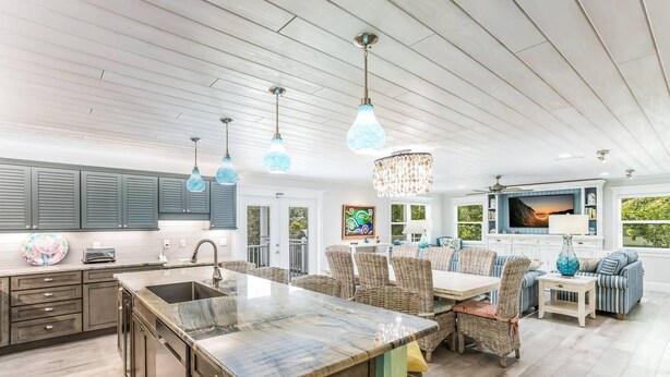 A spacious kitchen with a large island, dark wooden cabinets, and a dining area with wicker furniture. - Octopus Cottage: 1.5 Blocks to Beach, Pool & Spa by Anna Maria Life Vacation Rentals (Holmes Beach)