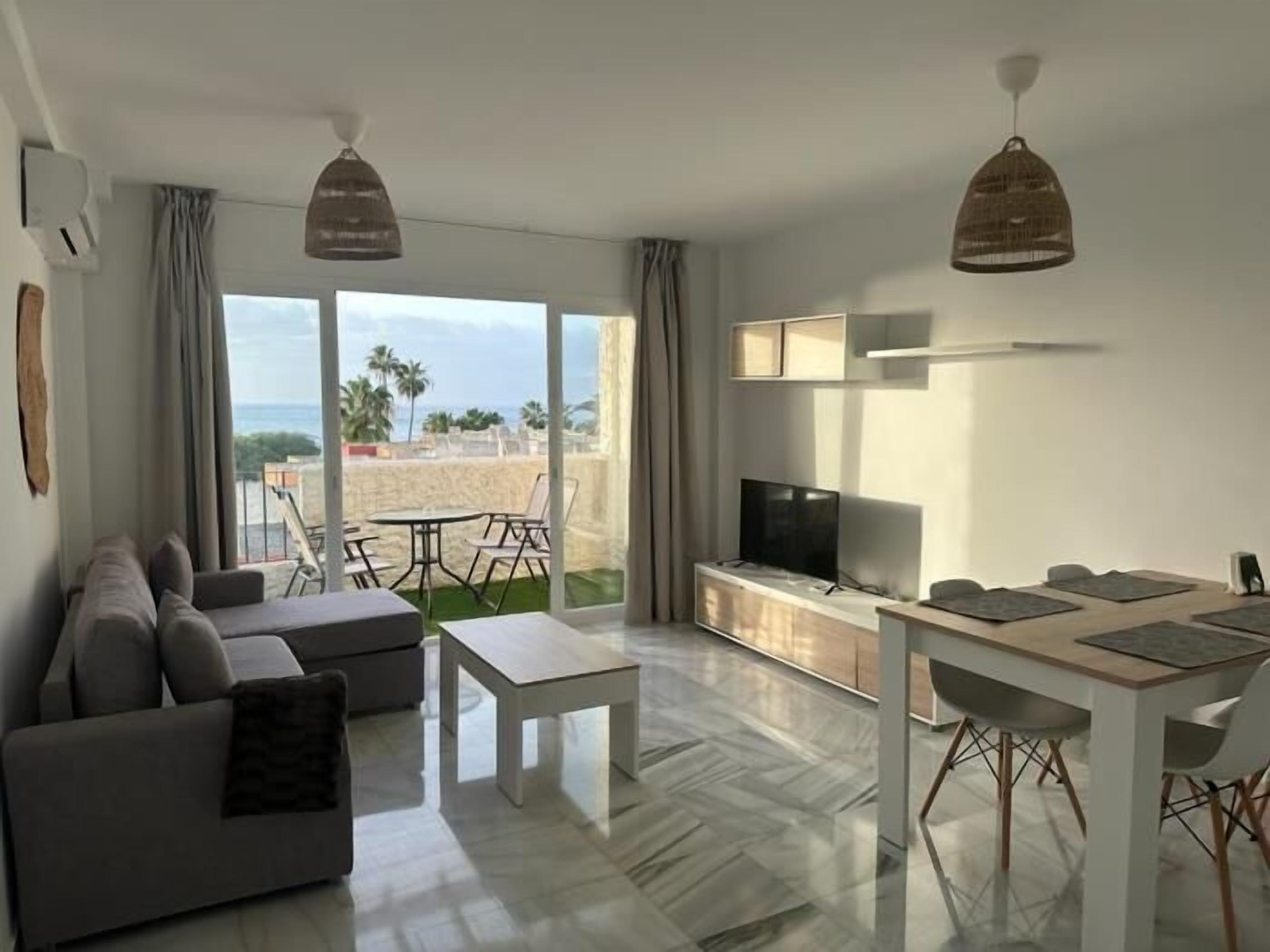 A modern living room with a sofa, coffee table, and dining table. There is a TV on a cabinet and a view of the beach through large windows.