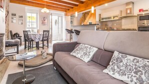 A cozy living room with a sofa, a small table, and a dog bed. There is a dining area with chairs and a table, and a kitchen in the background.