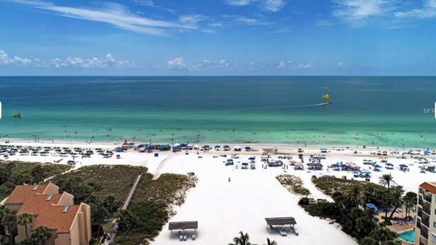 A beach with white sand, clear blue water, and a few umbrellas. There are buildings in the background.