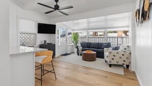 A spacious living area with a ceiling fan, a dark-colored sofa, a patterned armchair, and a dining area with a white table and wicker chairs.