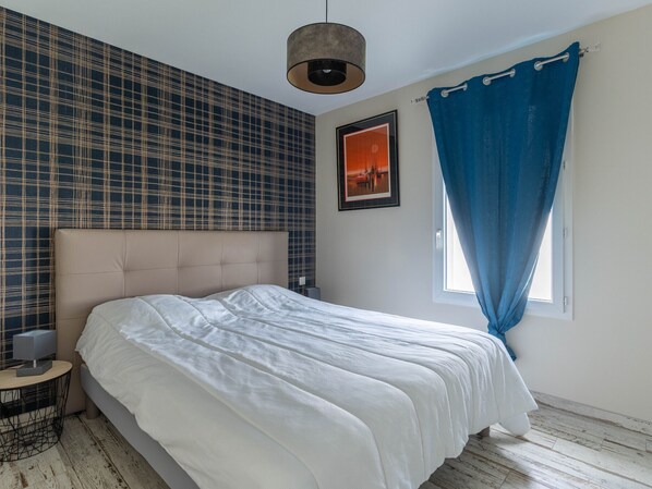 A bedroom with a large bed, a headboard, a bedside table with a lamp, a window with blue curtains, and a wall with a checkered pattern. - Cozy 3-Bed Home in Calm Parentis-en-Born, AC, Garden, Near Lake & Cycle Paths, Cleaning Included (Parentis-en-Born)