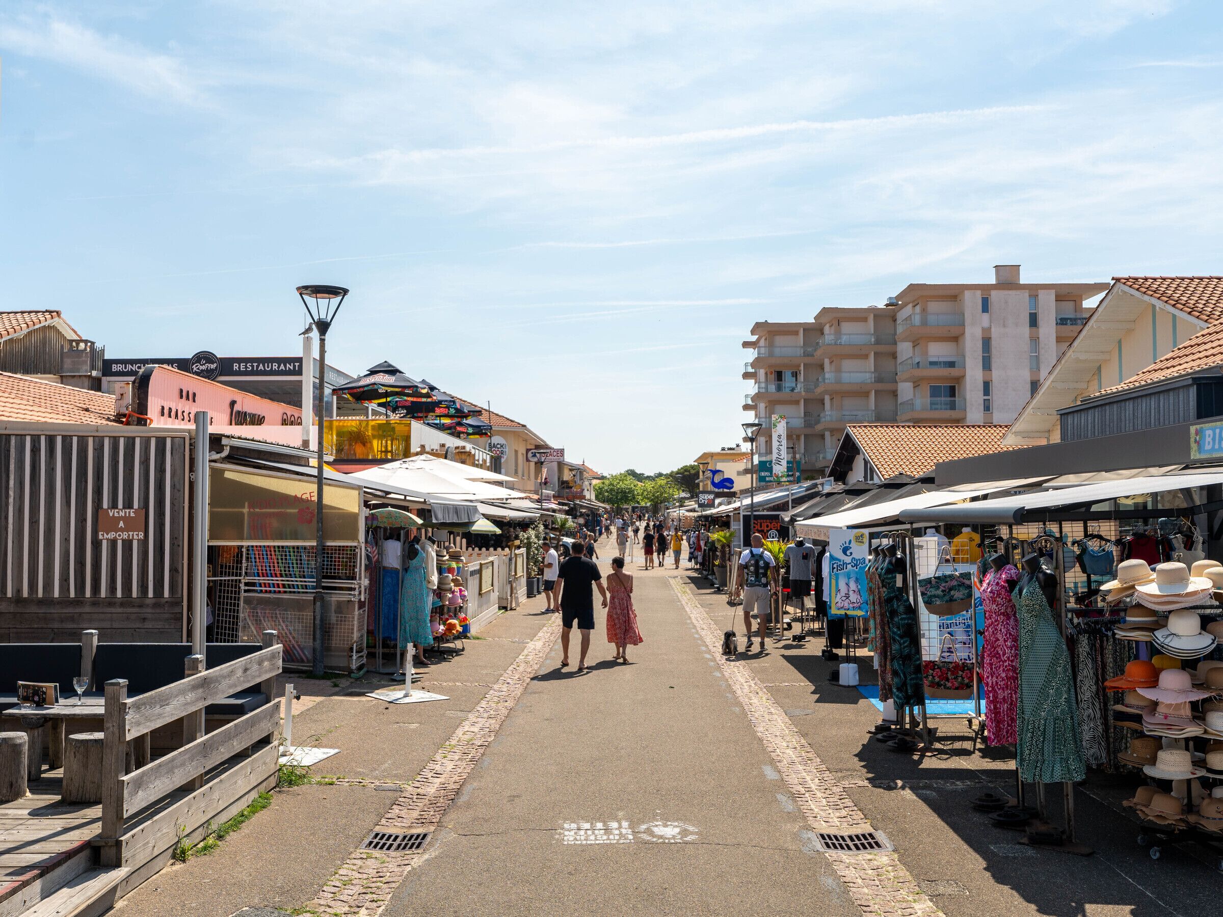 Un marché de rue animé, avec de nombreux étals et boutiques, un ciel dégagé et des bâtiments en arrière-plan.