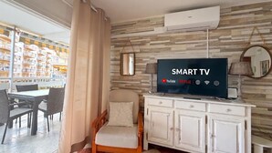 A hotel room with a TV displaying 'SMART TV' and streaming services, a wooden chair, a white cabinet, and a view of a balcony with outdoor seating.