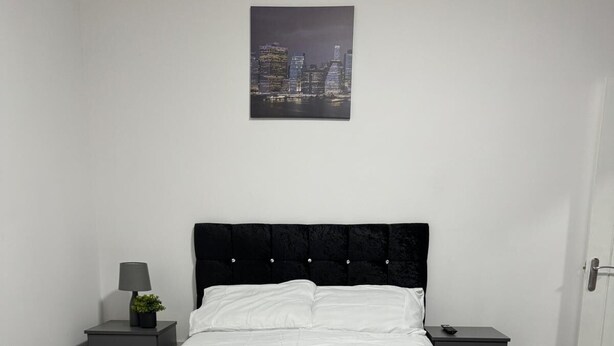 A neatly arranged bedroom with a bed, bedside tables, a lamp, and a cityscape painting on the wall. - 1-bed Apart-nottingham-free Parking (Nottingham)
