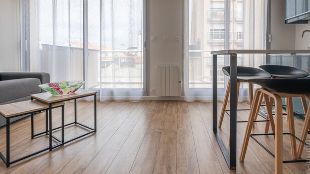 Een modern appartement met een houten vloer, een grijze bank, een klein tafeltje met een schaal, een eethoek met twee stoelen en grote ramen met gordijnen.