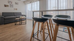 Een moderne woonkamer met een grijze bank, een klein tafeltje en twee zwarte stoelen. Er zijn grote ramen met dunne gordijnen en uitzicht op het omliggende landschap.