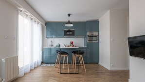 Een moderne keuken met blauwe kastjes, een wit aanrecht en een ingebouwde oven. Er staan twee krukken en een eettafel.