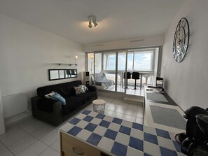 Uma sala de estar moderna com um sofá preto, uma mesa de centro xadrez e uma grande janela com vista para a praia.