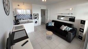 A compact living space with a kitchenette, a black sofa, a small round table, and a TV.