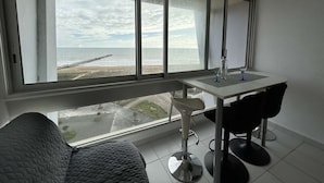 A room with a large window overlooking a beach, a small table with two chairs, and a bed with a quilted cover.