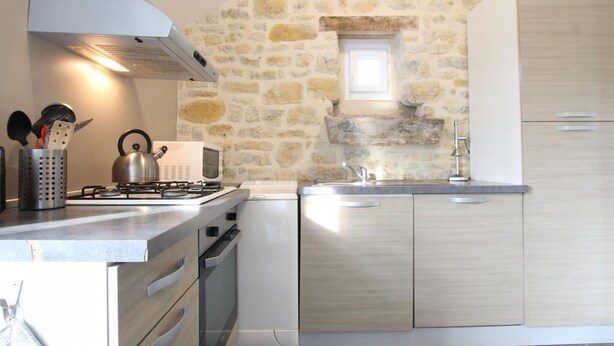 A modern kitchen with a stone wall, a gas stove, a microwave, and wooden cabinets. - Charming house near Utah Beach, pets allowed (Sainte-Marie-du-Mont)