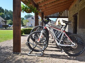 A covered bike parking area with a gravel ground, a wooden pergola, and a stone building in the background.