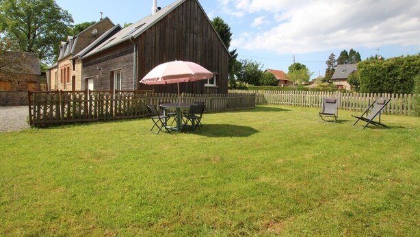 Une grange rustique avec une clôture en bois, une cour verdoyante et une table entourée de chaises, le tout abrité sous un parasol rose.