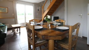 Un coin repas avec une table en bois dressée pour quatre personnes, entourée de chaises. On aperçoit un escalier au fond, qui mène à l’étage supérieur.