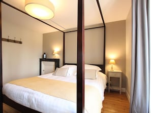 A bedroom with a four-poster bed, two bedside tables with lamps, a wooden floor, and a plain wall.