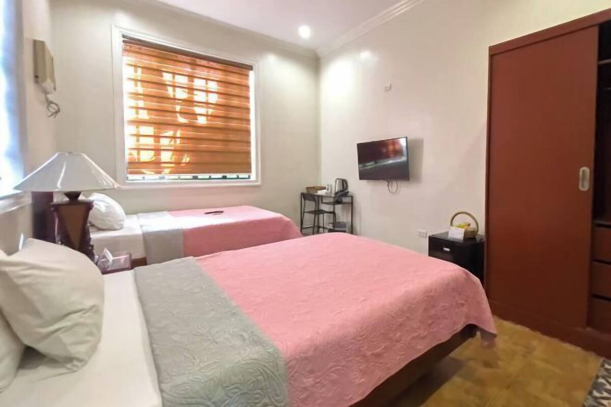 A bedroom with two beds, a TV, a desk, and a window with blinds.