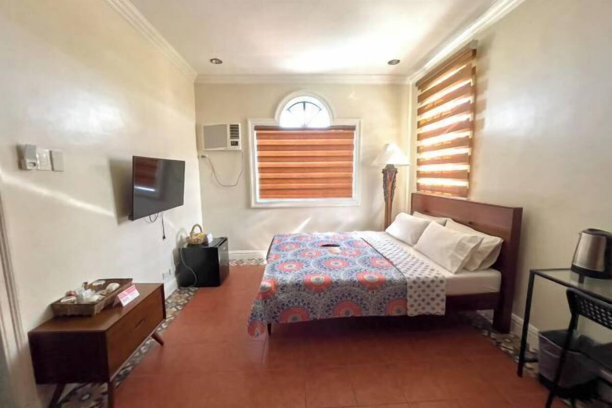 A hotel room with a bed, a TV, a window with blinds, a bedside table, and a lamp.