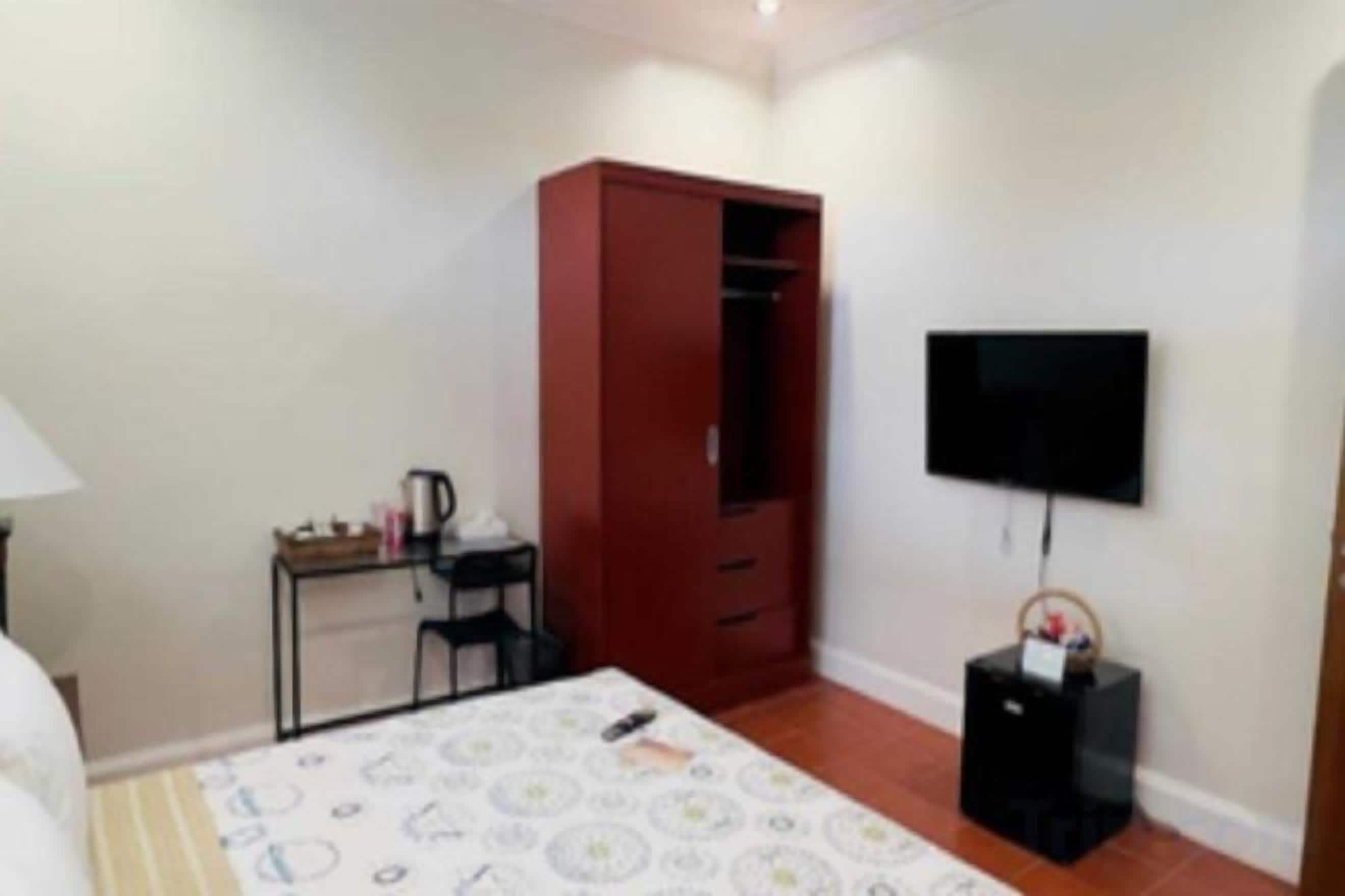 A bedroom with a bed, a wardrobe, a desk with a chair, a television, and a small table.