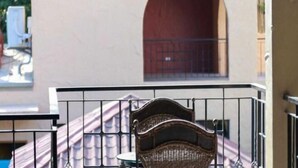 Un balcón con una silla de mimbre, baranda metálica y un edificio con puerta roja al fondo.