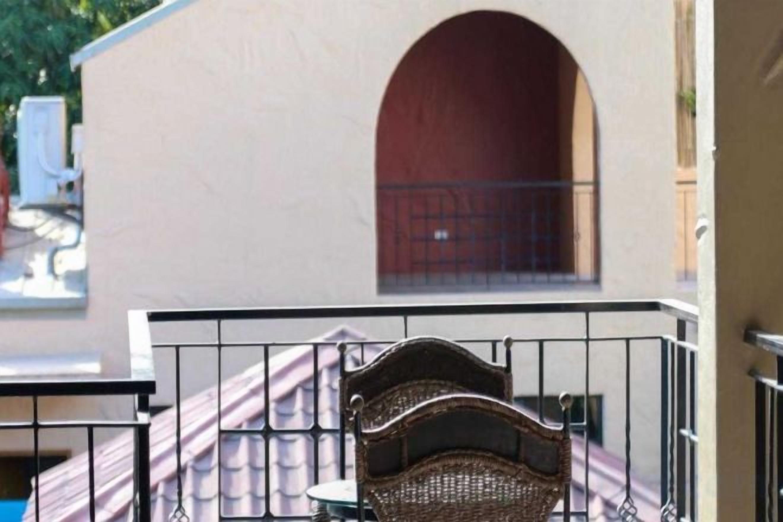 Un balcó amb una cadira de vímet i una taula, una paret blanca i una porta vermella al fons.