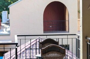 Un balcón con una silla de mimbre y una mesa, una pared blanca y una puerta roja al fondo.