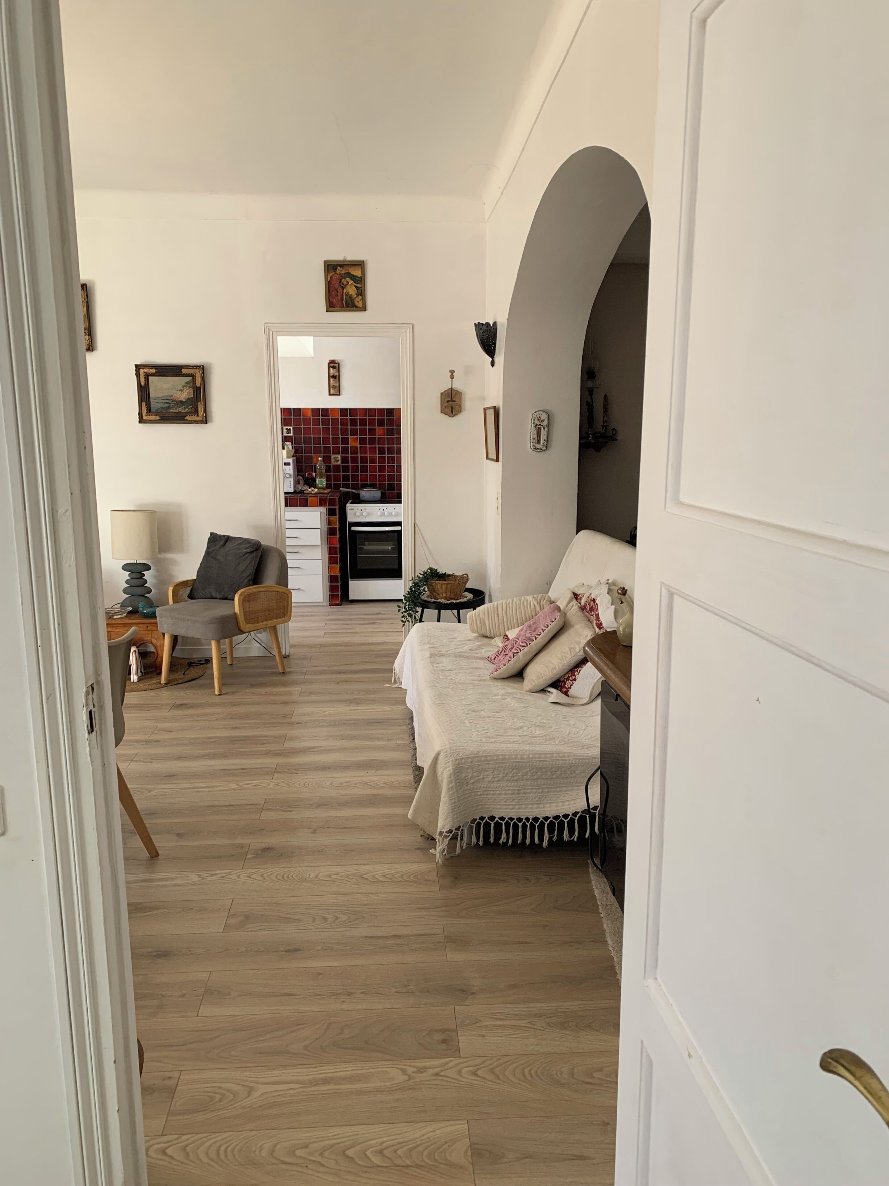 A room with a bed, a desk, a chair, and a kitchen area in the background.