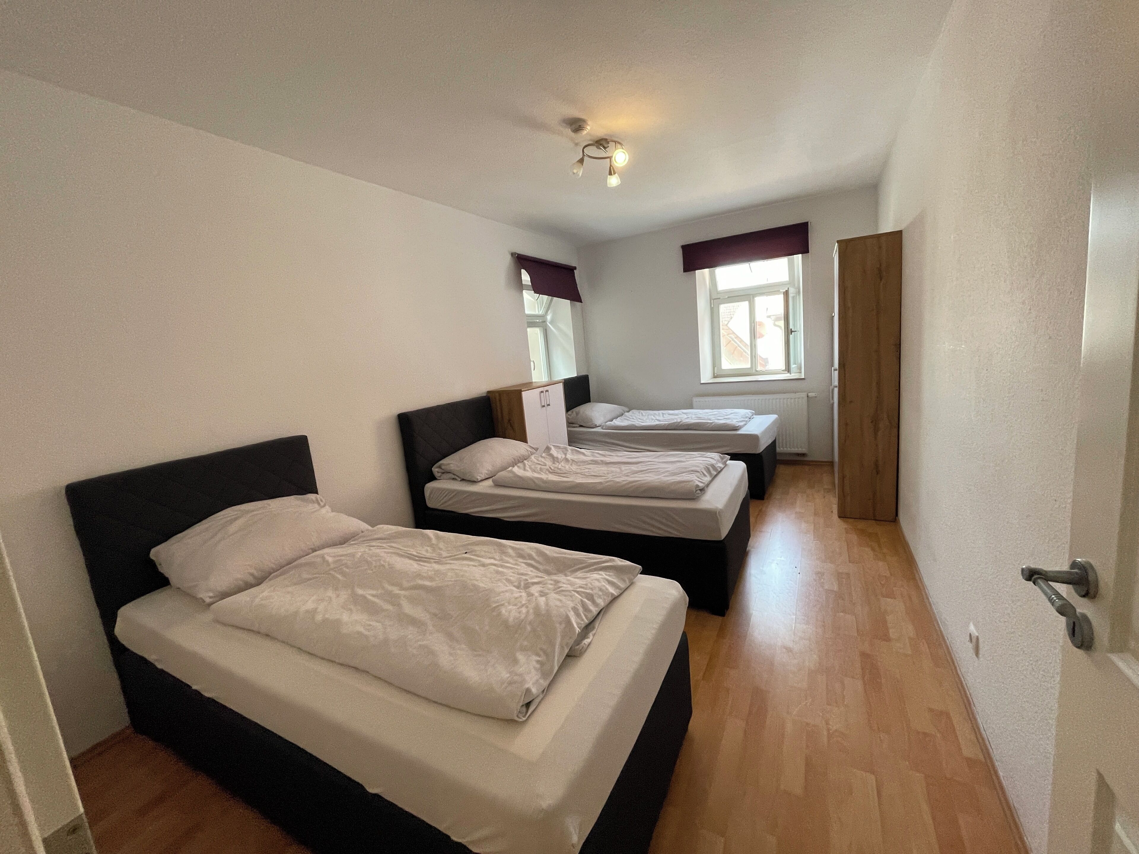 A small, clean room with three beds, a wooden wardrobe, and a window with curtains.