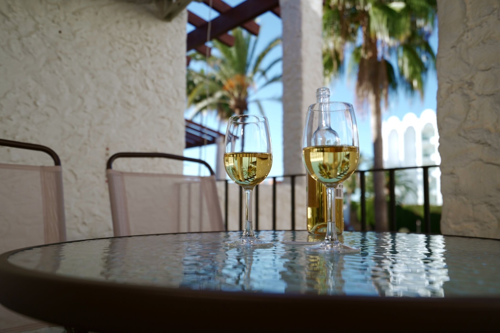 Two glasses of white wine on a round glass table with a view of palm trees and a white fence in the background.