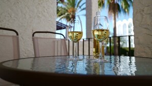 Two glasses of white wine on a round glass table with a view of palm trees and a white fence in the background.