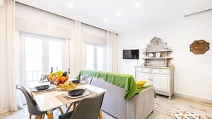 Uma área de jantar com uma mesa posta para dois, um sofá, uma televisão sobre um móvel e um tapete estampado.