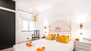 Quarto de hotel com uma cama grande, uma mesa de cabeceira com abajur, uma cadeira e uma janela com cortinas.