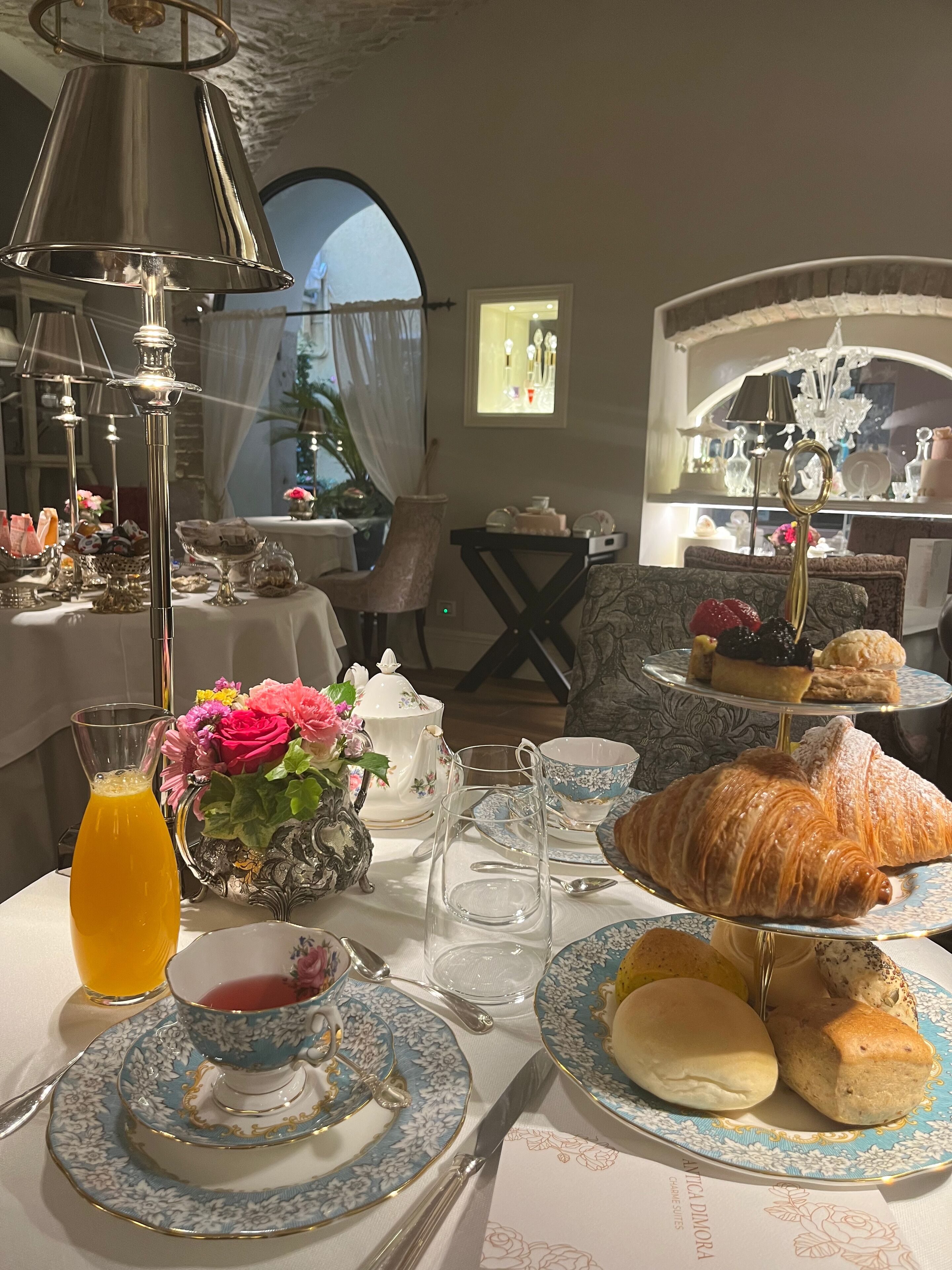 A breakfast setting with a table set for two, featuring a variety of pastries, a pitcher of orange juice, and a floral arrangement.