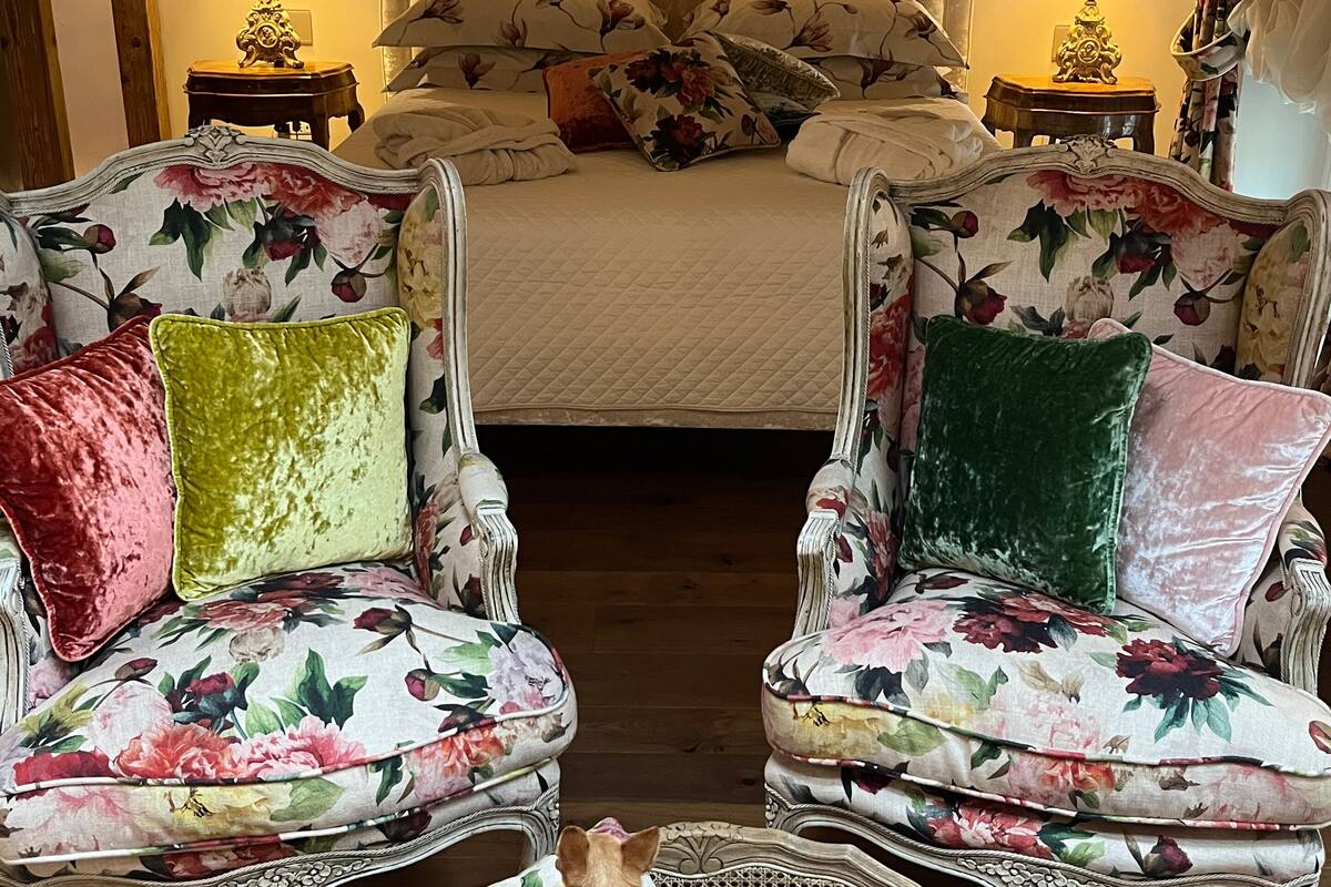 Un dormitorio con una cama, dos sillones tapizados con motivos florales, una pequeña cesta de mimbre con un perro y suelo de madera.