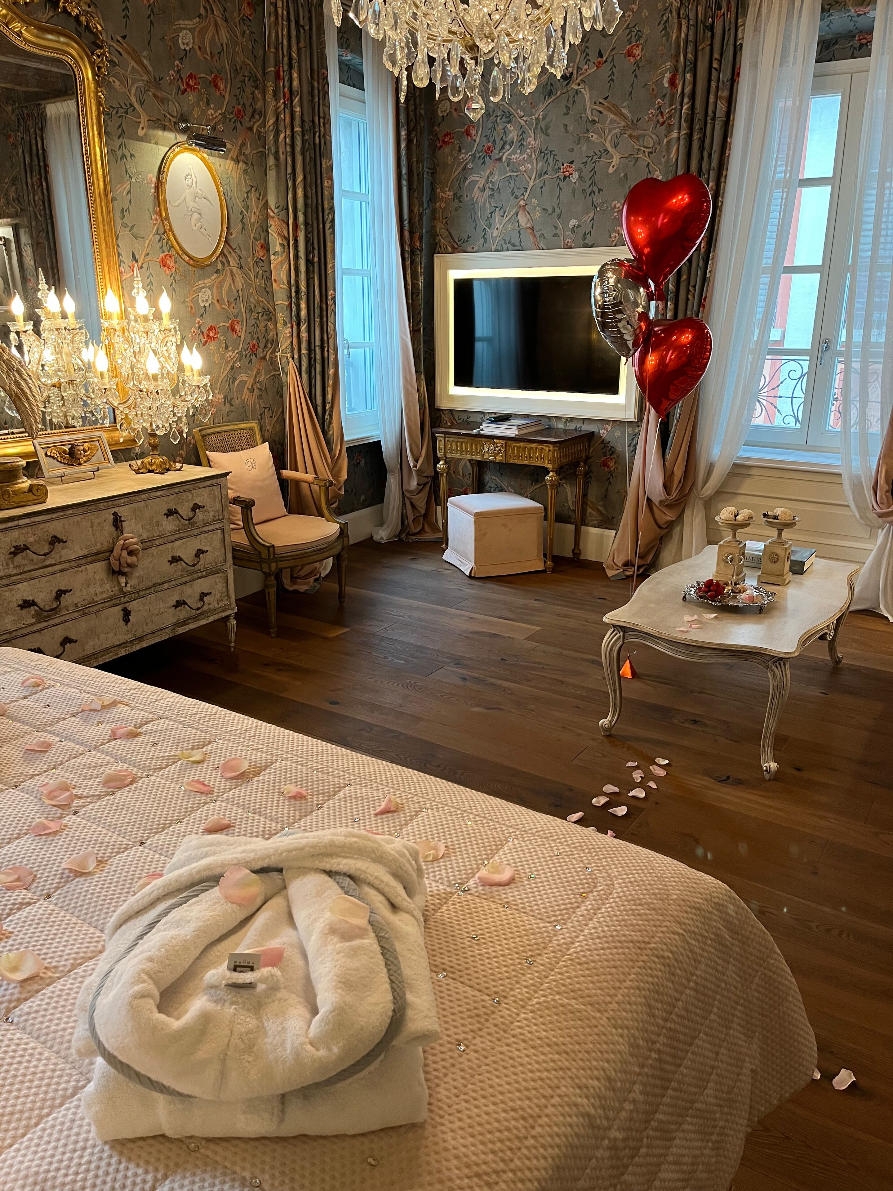 Ein luxuriöses Schlafzimmer mit einem großen Bett, einem Schminktisch mit Spiegel, einem Fernseher und einem kleinen Tisch mit herzförmigen Ballons.
