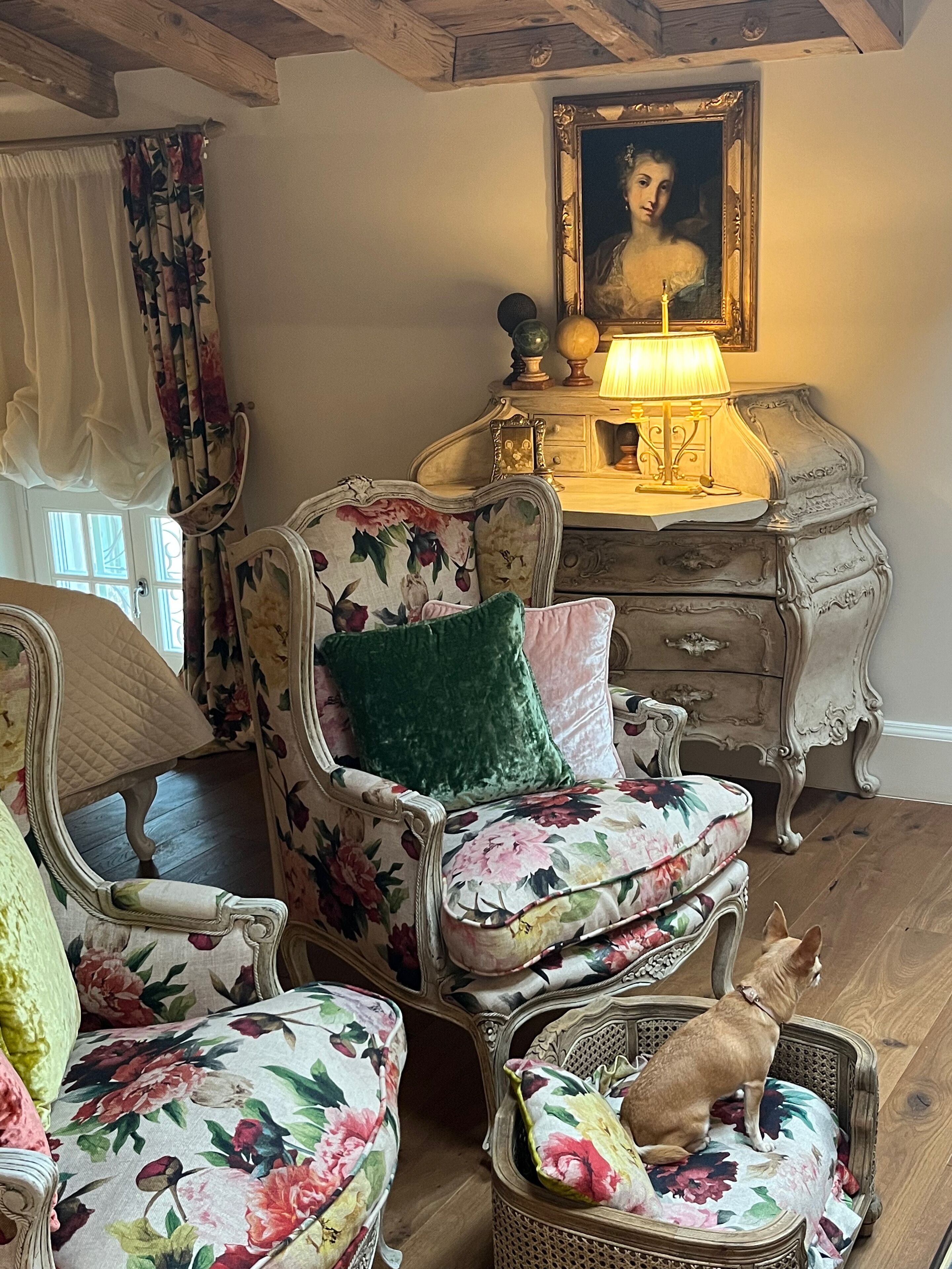 Ein Zimmer mit einem blumigen Sessel, einer Vintage-Kommode und einem gerahmten Porträt an der Wand.