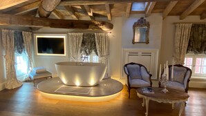 Une pièce avec une grande baignoire ronde en métal placée au centre, des poutres en bois au plafond, une grande fenêtre avec des rideaux et deux fauteuils rembourrés.