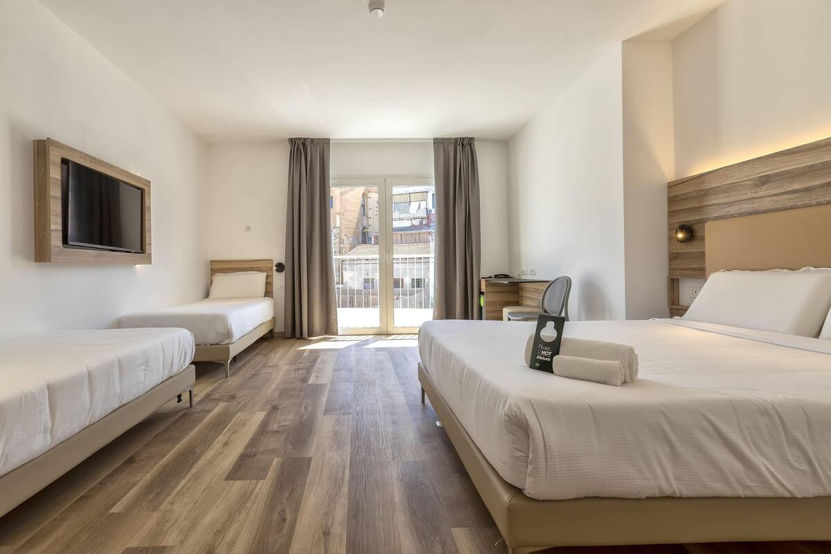 Camera d'albergo con due letti singoli, un letto matrimoniale, una TV a schermo piatto montata a parete, una scrivania con sedia e vista all'esterno tramite una porta vetrata.