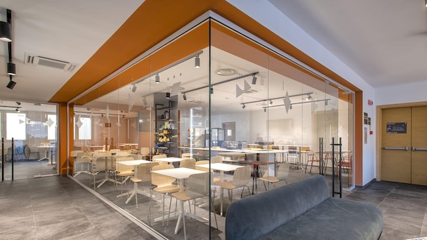 Un espacio de oficina moderno con paredes de vidrio, un área de comedor con mesas y sillas, y un sofá gris.