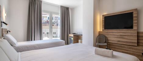 A hotel room with two beds, a TV mounted on the wall, a desk with a chair, and a view of the city through large windows.
