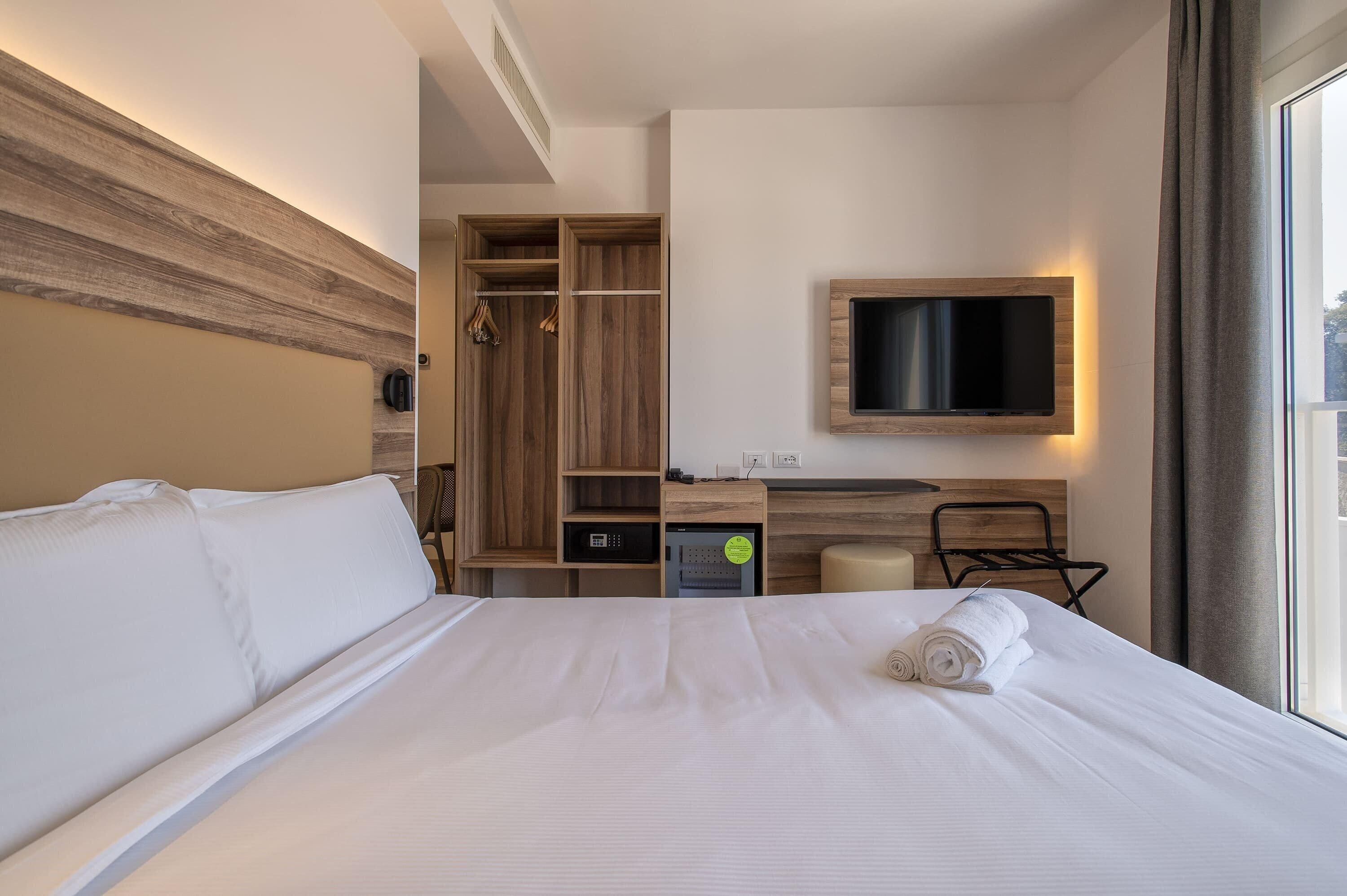 A hotel room with a large bed, a desk with a flat-screen TV, a wardrobe, and a window with curtains.