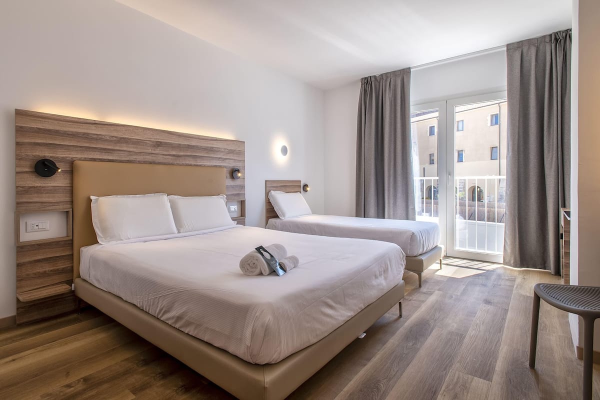 Camera d'albergo con due letti, una testiera in legno, un piccolo tavolo e vista sul balcone esterno.
