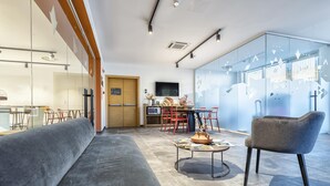 Una sala moderna con un sofá, una mesa de centro y una silla. Hay una zona de cocina con un televisor y sillas rojas.