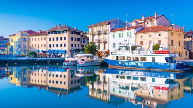 Ein Uferbild mit einem blauen Boot namens 'NUOVA CRISTINA', das in einer Marina vor Anker liegt, umgeben von Gebäuden mit Balkonen und roten Ziegeldächern.