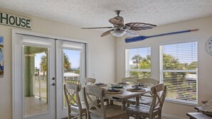 Zona pranzo con tavolo apparecchiato per quattro, sedie in legno, ventilatore a soffitto e vista sull'esterno attraverso le finestre.