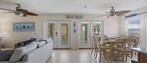 Ein wohnzimmerähnlicher Raum im Strandhausstil mit einer Couch, einem Esstisch, Stühlen, Deckenventilatoren und einem Schild mit der Aufschrift 'BEACH HOUSE'.