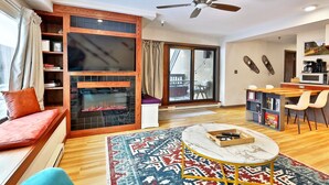 A modern living room with a fireplace, a ceiling fan, a dining area with chairs, and a view through the glass door. - Mountain Views at Mt Green: 2 BR/2BA, Shuttle, Discounted Ski Rentals, Pool, Hot Tub and More! (Killington)